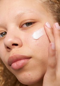 Gros plan sur un visage appliquant une crème blanche sur la joue. La peau est claire avec de légères imperfections ; les traits comprennent des yeux verts et des cheveux clairs et bouclés.