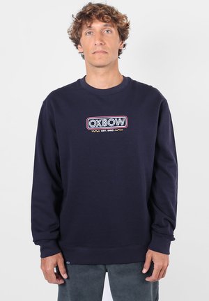Homme aux cheveux bouclés portant un sweat-shirt bleu marine avec le logo "OXBOW EST. 1985" imprimé sur la poitrine, debout devant un fond uni.