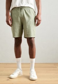 Lichtgroene shorts van textiel, met een elastische tailleband en een trekkoord, en zijzakken. Draagt witte sneakers en sokken.