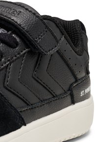 Zapatilla negra que presenta materiales de cuero y ante, una correa de gancho y bucle, acentos en zigzag y una suela de goma blanca etiquetada "ST. POWER".