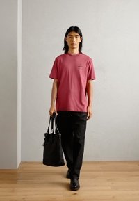 Tricou roz, cu mânecă scurtă și decolteu rotund, purtat împreună cu pantaloni negri și o geantă neagră, pe o persoană care stă în interior.