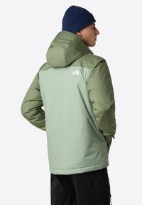 Vihreä eristetty takki, jossa on tummempi vihreä yläosa, huppu ja vetoketjulliset taskut. Takissa on logo olkapäällä ja teksturoitua kangasta.