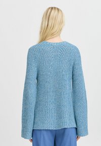 Femme blonde portant un pull en maille texturée bleu et un pantalon bleu assorti, debout de dos contre un fond uni.
