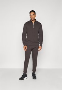 Mørkebrun hettejakke med glidelås og lange ermer, ribbestrikkede ermer, og matchende smale bukser, kombinert med svarte joggesko.