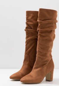 Bottes en suede marron, hautes jusqu'au genou, au design effilé, avec un bout pointu et un talon bloc de hauteur moyenne. Texture lisse avec un subtil éclat.