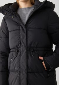 Giacca a vento nera con cappuccio, caratterizzata da un design trapuntato, vita regolabile con cordino, tasche con zip e un tessuto morbido e strutturato.