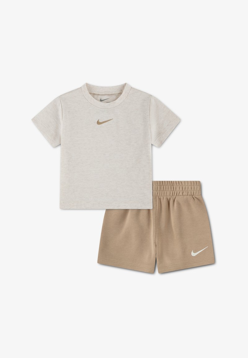 Bež kratka majica z rokavi iz blagovne znamke Nike z zlatim logotipom, kombinirana s svetlo rjavimi elastičnimi kratkimi hlačami Nike z belim logotipom na levi nogi.