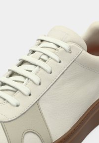 Ανδρικό αθλητικό παπούτσι από κρεμ δέρμα με ανοιχτό γκρι λεπτομέρειες, επίπεδα κορδόνια και υφή στο επάνω μέρος. Διαθέτει στρογγυλεμένο σχήμα και ευέλικτη καφέ ελαστική σόλα.