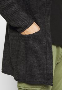 Cardigan nero con trama, dal taglio ampio, con una grande tasca e polsini a coste, abbinato a pantaloni verde oliva.