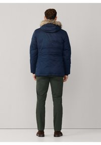 Chaqueta tipo puff azul marino con capucha forrada de piel, combinada con pantalones verdes. La chaqueta presenta un ligero brillo y un corte más largo.