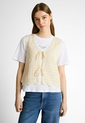 Junge Frau trägt ein weißes T-Shirt unter einer cremefarbenen Strickweste mit Schnüren, kombiniert mit blauen Jeans, steht vor weißem Hintergrund.