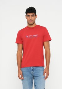Camiseta de algodón roja con mangas cortas, que presenta un gráfico del logo azul "tommy" en el pecho. Combinada con jeans de denim azul claro.