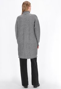 Szary, dziany długi kardigan z wysokim kołnierzem, teksturowym wzorem w zygzaki na plecach oraz prążkowanym dołem. Nosi się go na czarnych spodniach.
