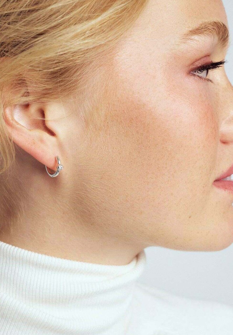 Blond kvinna i vit polotröja som visar en liten silverhänge med en enda ädelsten i sitt vänstra öra, profilen synlig.