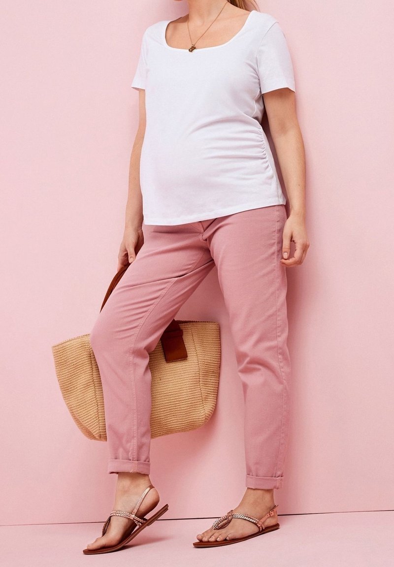 Top blanco de manga corta y escote cuadrado combinado con pantalones entallados rosas. La persona sostiene un bolso de yute marrón y lleva sandalias planas con adornos decorativos.