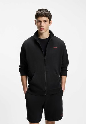 Ung mand iført en sort lynlåstrøje og matchende shorts med røde "HUGO"-logoer på brystet og benet, står med hænderne i lommerne.