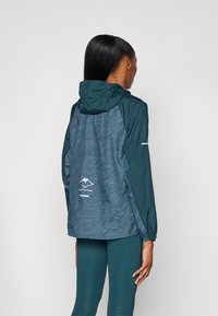 Teal vandtæt jakke med en tekstureret, mønstret ryg, hætte og reflekterende detaljer. Har logo tryk og elastiske manchetter.