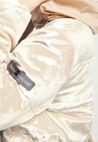 Chaqueta acolchada de color beige claro, fabricada en un tejido suave y brillante con textura acolchada. Presenta una etiqueta con el logo en la manga y puños ajustados.