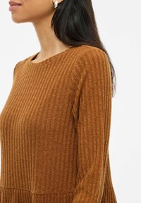Robe en tricot marron côtelé avec un col rond et des manches longues. Le tissu présente un motif texturé et une ourlet en volants.
