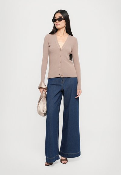 Cardigan beige a coste con scollo a V e chiusura a bottoni, abbinato a jeans a gamba larga in denim scuro e una borsa in metallo argentato. Modella che indossa occhiali da sole.