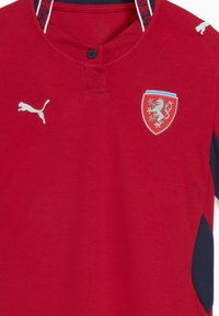 Maillot de sport rouge avec un col boutonné, arborant un logo Puma blanc et un emblème en forme de bouclier avec un dessin de lion. Panneaux latéraux bleu foncé.
