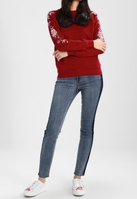 Sudadera roja con bordado floral en las mangas, combinada con jeans ajustados de mezclilla oscura con franjas laterales, y zapatillas blancas.
