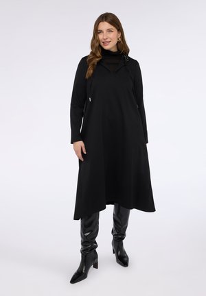 Vestido negro de manga larga con cuello alto y dobladillo asimétrico, con una superposición texturizada y tachuelas en el cuello. Llevado con botas ajustadas negras.