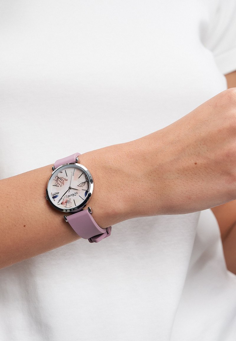 Montre ronde avec un boîtier argenté, un bracelet en silicone rose clair, et un cadran blanc à motif floral avec des index et des aiguilles argentés.