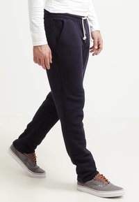 Marinblå sweatpants med elastiskt midjeband och snörning, med sidofickor och en avsmalnad passform, tillsammans med grå sneakers.