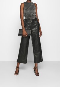 Top senza maniche grigio metallico con arricciature, abbinato a pantaloni larghi in pelle nera. Tiene una pochette nera e indossa décolleté nere con lacci.