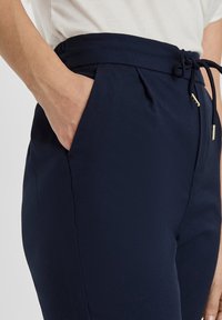 Granatowe, zwężane spodnie uszyte z teksturowanej tkaniny, z elastycznym paskiem w talii i sznurkiem do regulacji oraz kieszeniami po bokach. Złote akcenty z metalu.