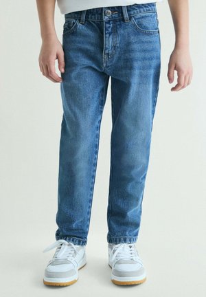 Niño con vaqueros azules y zapatillas blancas de pie contra un fondo liso, con las manos relajadas a los lados.