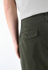 Close-up of a person wearing dark green pants with a buttoned back pocket and a tucked white shirt, showing part of their arm and waist.