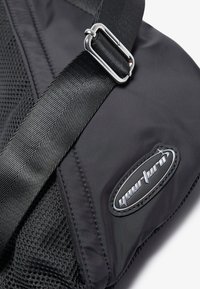 Borsa a tracolla nera con pannello in rete testurizzata, tracolla robusta e accessori in metallo argentato. Patcher con logo visibile su un lato. Superficie in tessuto liscia.