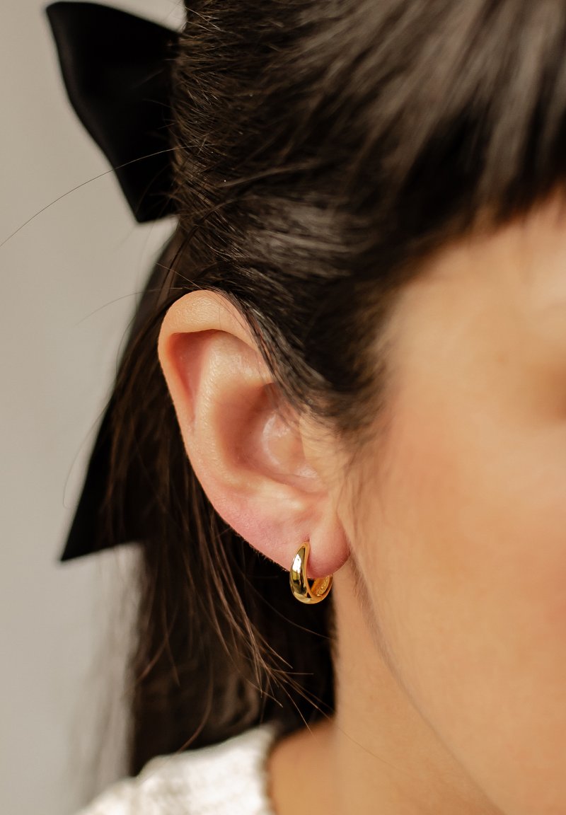 Boucle d'oreille en or avec une finition lisse, présentée en gros plan sur une oreille. Les cheveux sont foncés et coiffés avec un ruban noir.