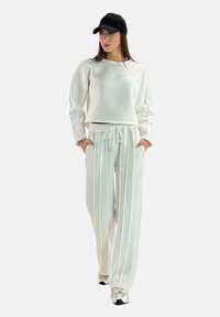 Sweat-shirt en polaire blanche avec manches surdimensionnées, associé à un pantalon à cordon de serrage assorti présentant des rayures verticales. Une casquette noire contraste avec la tenue.