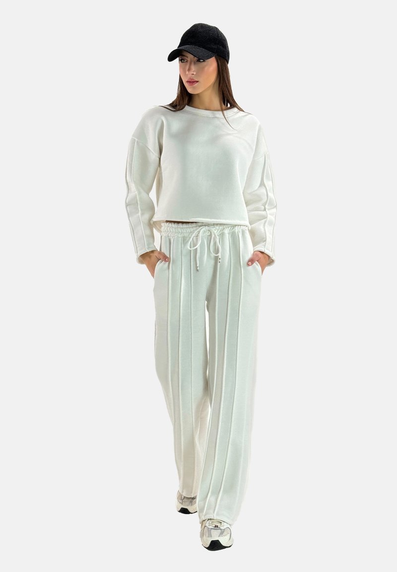 Witte, zachte stoffen trainingspak bestaande uit een cropped, boxy sweatshirt en hoog- taille, wijde broek met verticale strepen en zakken. Zwarte pet.