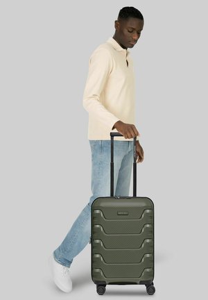 Homme en chemise beige à manches longues et jeans tenant la poignée d'une valise rigide verte à roulettes sur fond uni.