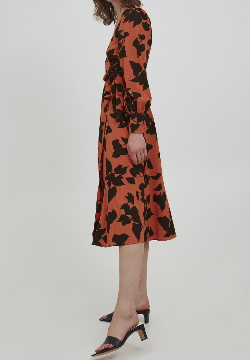 Robe en tissu texturé orange avec un motif de feuilles noires, comportant des manches longues, une taille cintrée à nouer et une jupe fluide. Sandales à talons noires.