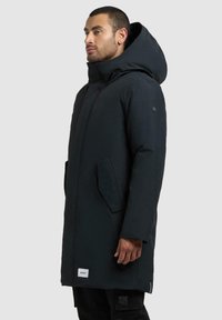 Czarny długi parka z kapturem, zapinana na zamek, z dwiema bocznymi kieszeniami i materiałową metką u dołu. Gładka tekstura, nowoczesny design.