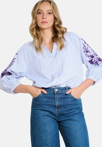 Femme portant un blouson rayé bleu et blanc avec des broderies florales violettes sur les manches et un jean bleu taille haute, les mains dans les poches.