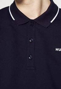Polo shirt bleu marine foncé avec un col à rayures blanches contrastées et une patte de boutonnage à trois boutons, ainsi qu'un petit logo blanc sur la poitrine. Tissu texturé.