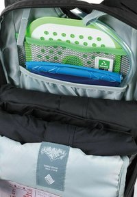 Zwarte rugzakinterieur met een grijze compartiment waarin een blauwe pen, een groene netzak en een logo-patch tegen een lichtblauwe stoffen achtergrond te zien zijn.