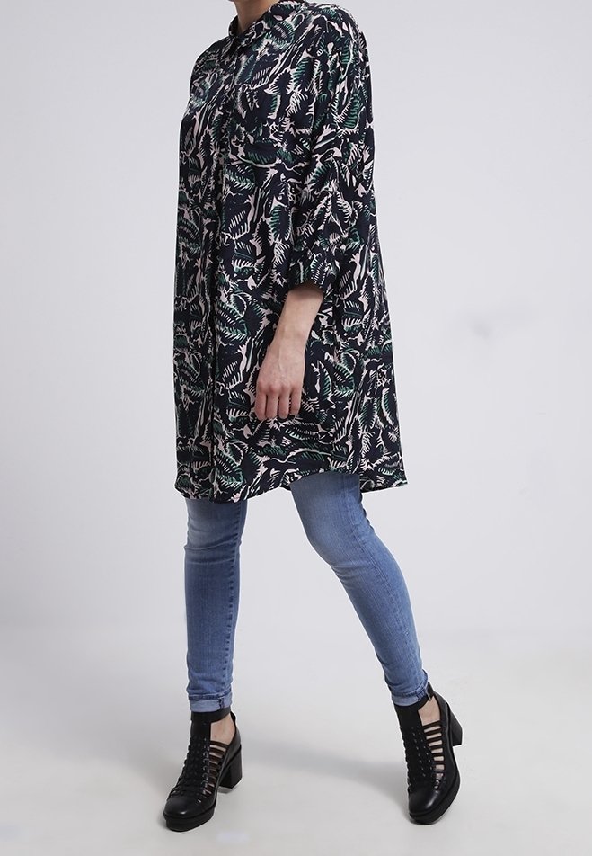 Personne portant une longue tunique à motifs floraux, un jean bleu ajusté et des bottines noires à talons lacées, debout les jambes croisées.