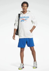Hvit hettegenser med blå Reebok-logo, kombinert med blå shorts. Modellen har på seg grå og gule joggesko og holder en svart ryggsekk.