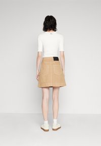 Top bianco aderente e gonna in denim khaki con due tasche posteriori. Presenta un'etichetta nera con il marchio sulla vita. Il modello indossa scarpe bianche con calze.