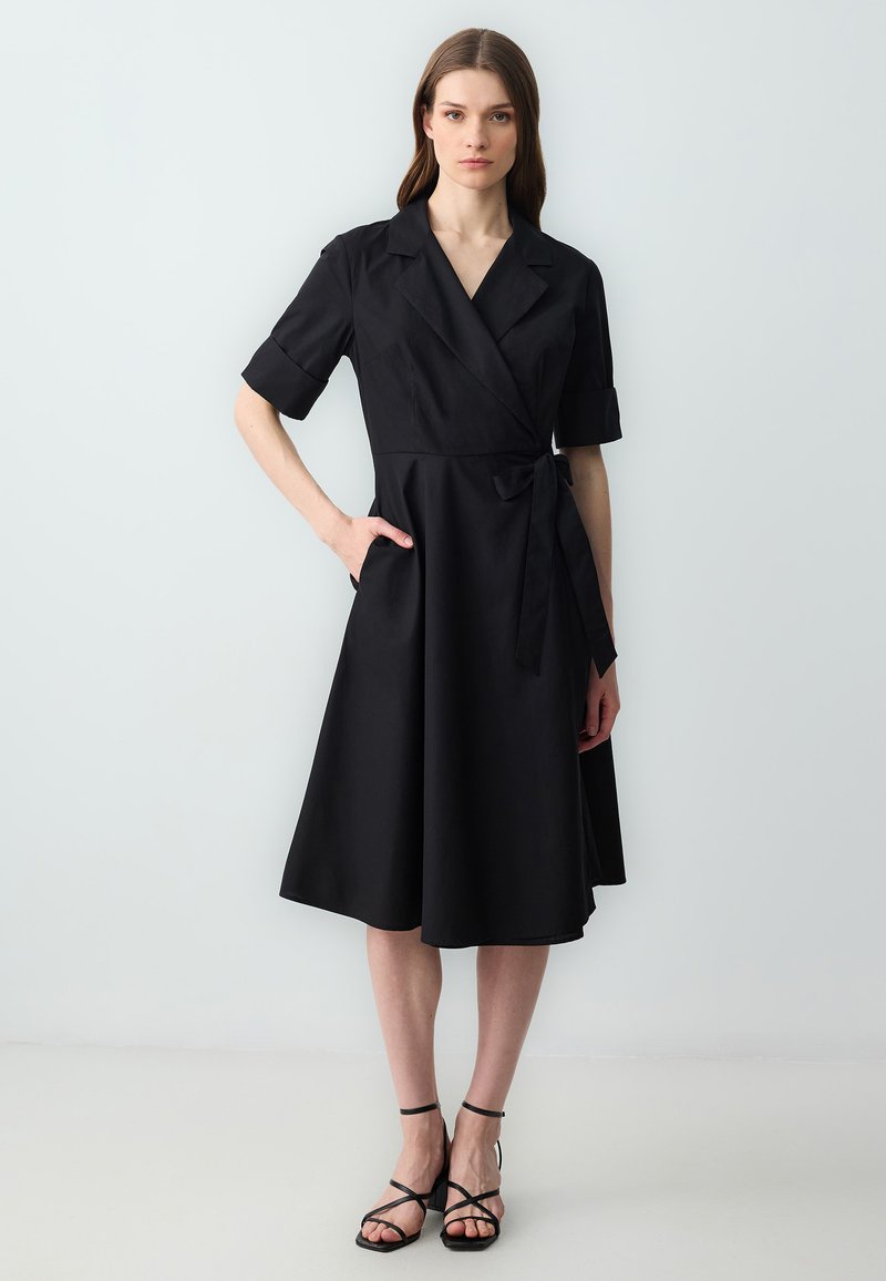 Robe noire portefeuille à manches courtes, avec un col, taille cintrée et poches latérales ; tissu lisse avec une jupe fluide.