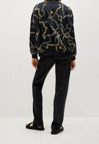 Chaqueta bomber de satén con fondo azul marino y patrón de cadenas doradas, combinada con jeans negros y sandalias negras, vista desde la parte posterior.