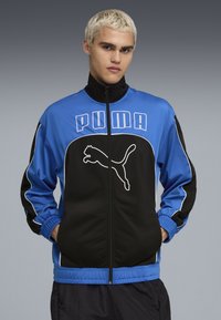 PUMA uzvelkamā jaka ar zilu un melnu krāsu shēmu, elastīgiem aizdariem un izteiksmīgu logotipa dizainu priekšpusē. Gluda tekstūra.