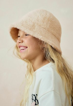 Un chapeau seau doux en pêche, fabriqué dans un matériau moelleux, avec un large bord et une couronne arrondie. Complété par des cheveux blonds clairs.